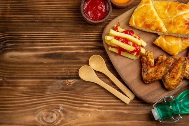 Vista superior de primer plano pastel de comida rápida y alitas de pollo papas fritas con salsa de tomate en el tablero de la cocina junto a los tazones de especias y salsas coloridas cucharas de madera hierbas y botella