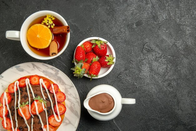 Vista superior de primer plano pastel de fresas con crema de chocolate con fresas y chocolate y una taza de té junto a los cuencos de fresas y crema de chocolate en el lado izquierdo de la mesa oscura