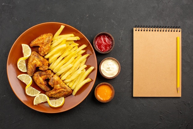 Vista superior de primer plano plato de comida rápida de alitas de pollo, papas fritas y limón junto a cuencos de tres tipos de salsas y cuaderno con lápiz sobre la mesa