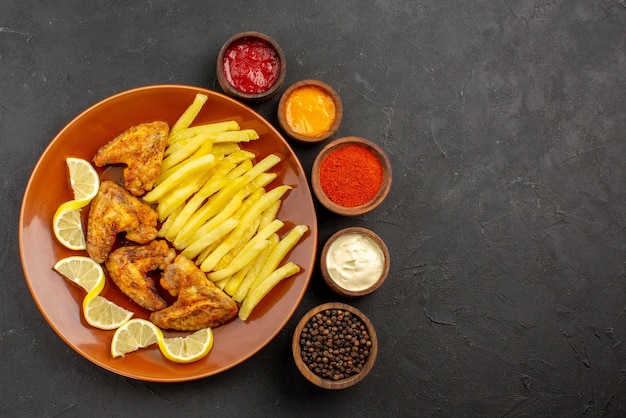 Vista superior de primer plano plato de comida rápida de alitas de pollo, papas fritas y limón y tazones de tres tipos de salsas, pimienta negra y especias en el lado izquierdo de la mesa