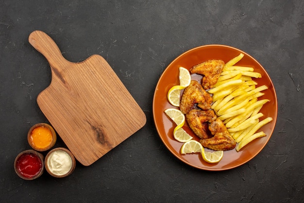 Foto gratuita vista superior de primer plano plato de comida rápida de unas apetitosas alitas de pollo, papas fritas y limón a la derecha y tres tipos de salsas junto a la tabla de cortar en el lado izquierdo