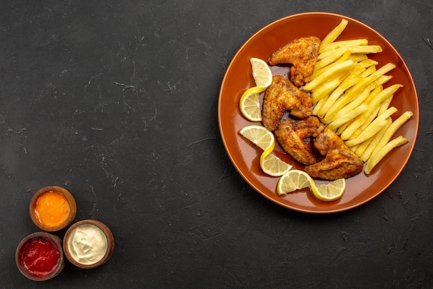 Vista superior de primer plano plato de comida rápida de un apetitoso alitas de pollo, papas fritas y limón a la derecha y tres tipos de salsas en el lado izquierdo de la mesa