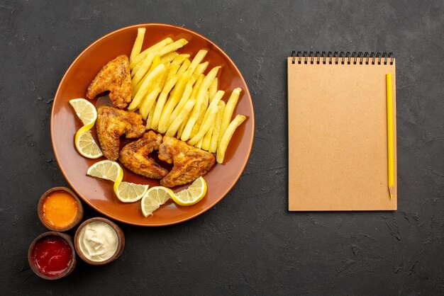 Vista superior de primer plano plato naranja de comida rápida de unas apetitosas alitas de pollo, papas fritas y limón con tres tipos de salsas junto al cuaderno de crema y lápiz sobre la superficie oscura