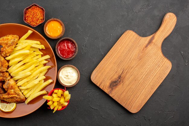 Vista superior de primer plano pollo y papas alitas de pollo papas fritas y limón tres tazones de diferentes tipos de salsas y especias junto a la tabla de cortar de madera en la mesa oscura