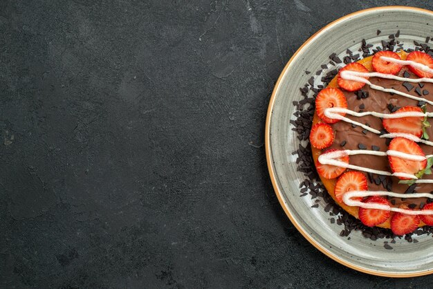 Vista superior de primer plano postre apetitoso pastel con trozos de fresa y chocolate en un plato blanco en el lado derecho de la mesa oscura