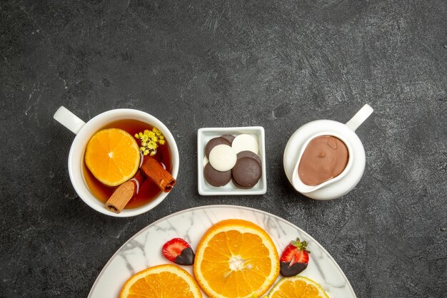 Vista superior de primer plano una taza de té plato de frutas cítricas y fresas junto a los tazones de chocolate y crema de chocolate y una taza de té con palitos de limón y canela