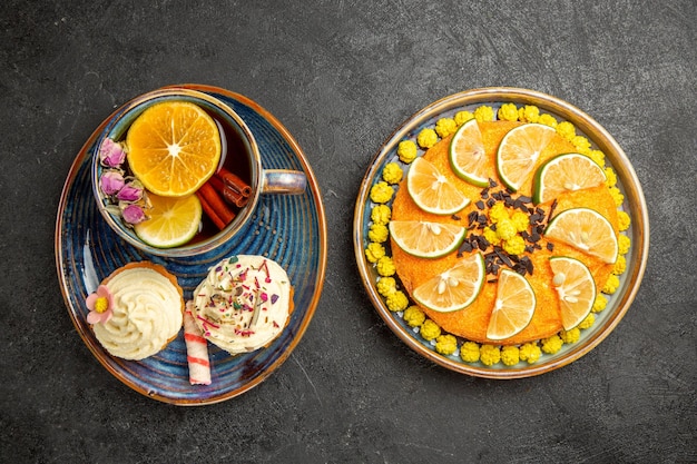 Vista superior de primer plano té de hierbas una taza de té de hierbas con limón y dos cupcakes con crema junto al plato de un apetitoso pastel con limas en la mesa negra