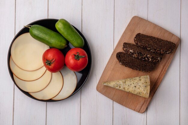 Vista superior de queso ahumado con tomates y pepinos en un plato con rebanadas de pan negro sobre un soporte sobre un fondo blanco.