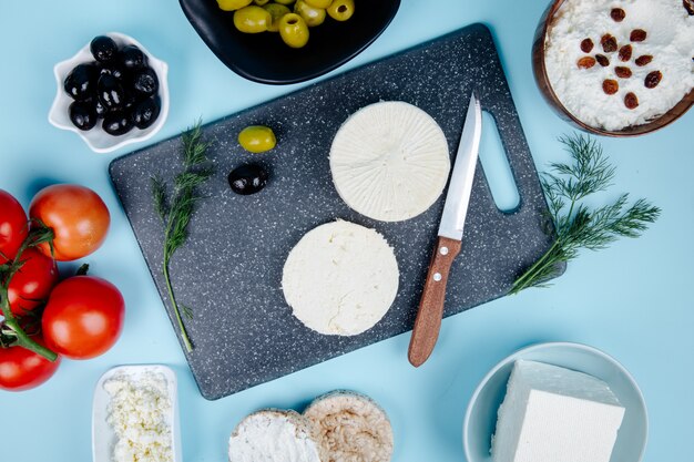 Vista superior de queso de cabra en un tablero negro con un cuchillo de cocina y tomates frescos aceitunas en vinagre y queso cottage en un recipiente en azul