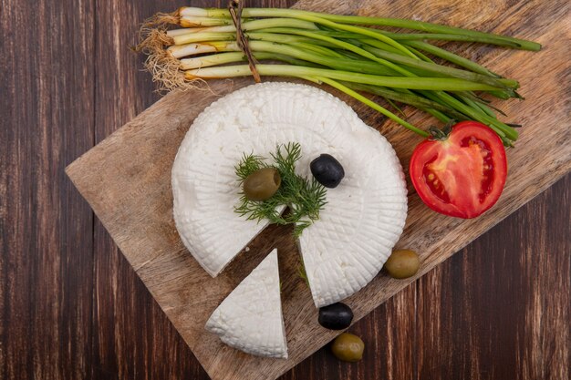 Vista superior de queso feta con aceitunas, tomate y cebollas verdes en un soporte sobre un fondo de madera