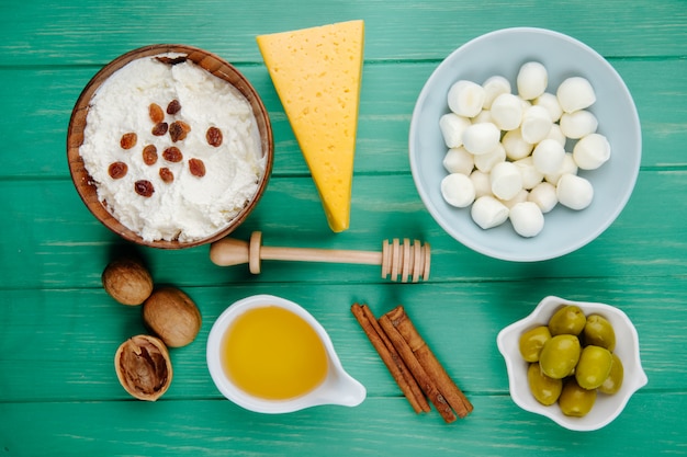 Vista superior de queso mozzarella en un tazón de requesón y un trozo de queso holandés con nueces palitos de canela y miel y aceitunas en escabeche en madera verde