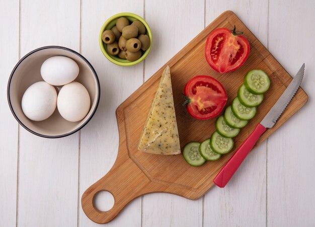 Vista superior de queso con tomate, pepino y aceitunas con un cuchillo en un soporte con aceitunas y huevos de gallina sobre un fondo blanco.