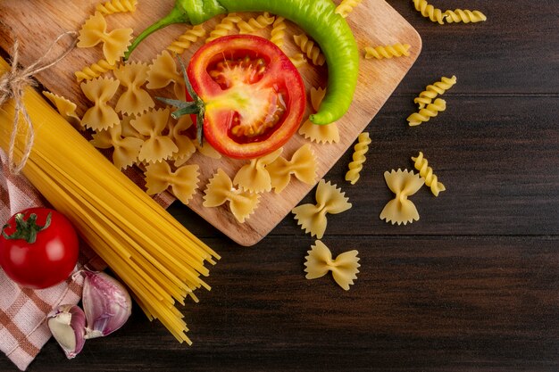 Vista superior de racimos de pasta cruda con ajo y pasta con tomate y pimiento picante en una pizarra sobre una superficie de madera