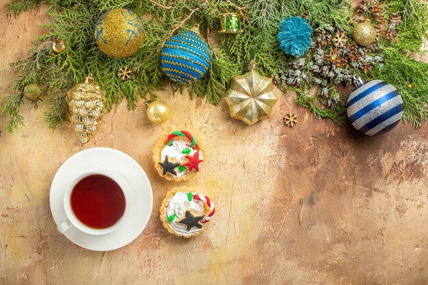 Vista superior de ramas de abeto adornos de árbol de Navidad una taza de té cupcakes sobre fondo beige