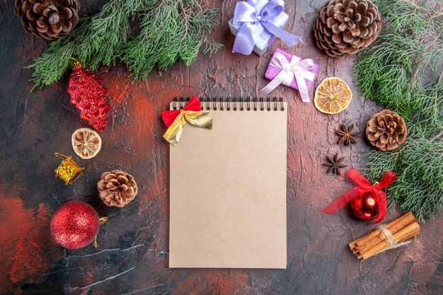 Vista superior de las ramas de los árboles de pino con conos anís canela regalos de navidad y colgantes un cuaderno sobre fondo rojo oscuro navidad