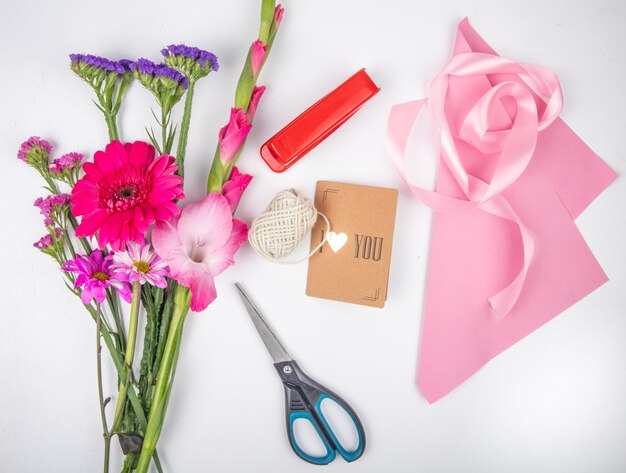 Vista superior de un ramo de flores de color rosa gerbera y gladiolo con statice y una grapadora roja con tijeras de cinta rosa y una pequeña postal sobre fondo blanco.