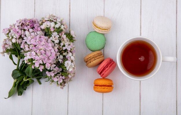 Vista superior del ramo de flores con una taza de té y macarons de colores sobre una superficie blanca