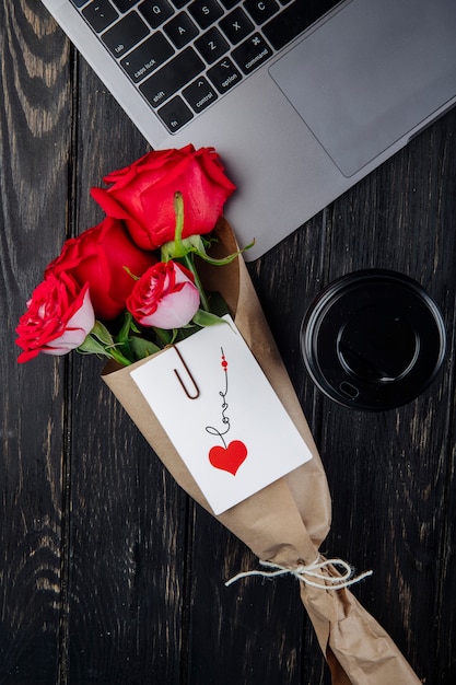 Vista superior de un ramo de rosas rojas en papel artesanal con postal adjunta acostada cerca de una computadora portátil con una taza de café de papel sobre fondo de madera oscura