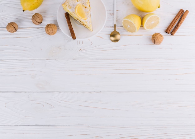 Foto gratuita vista superior de la rebanada de pastel decorado; limón; nueces canela; cuchara sobre fondo de madera blanco