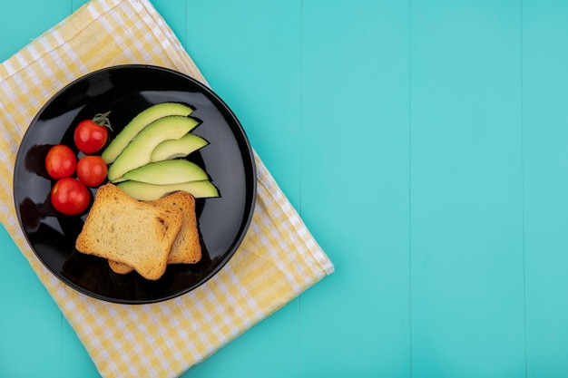 Foto gratuita vista superior de rebanadas de aguacate con tomates rebanadas de pan tostado en placa negra sobre mantel a cuadros amarillo sobre azul