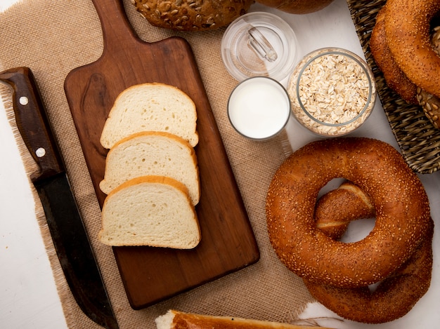 Vista superior de rebanadas de pan blanco en la tabla de cortar con bagels de leche con cuchillo y copos de avena sobre fondo blanco.