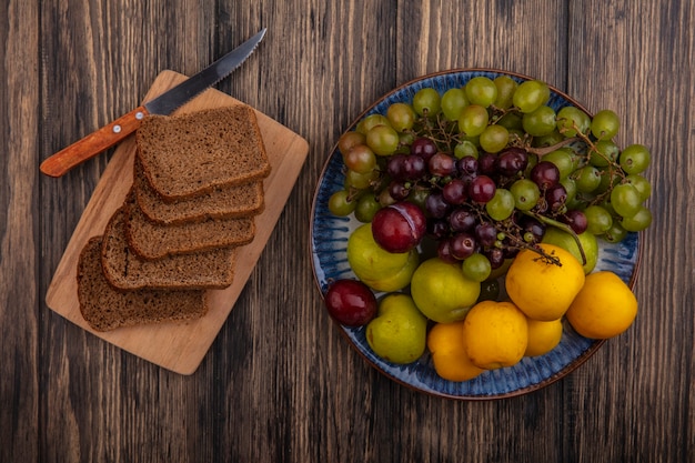 Foto gratuita vista superior de las rebanadas de pan de centeno y un cuchillo en la tabla de cortar con una canasta de uvas nectacots pluots sobre fondo de madera