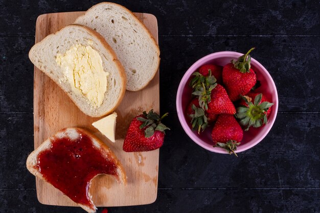 Vista superior rebanadas de pan y mantequilla con una rebanada de pan con mermelada a bordo con fresas en taza