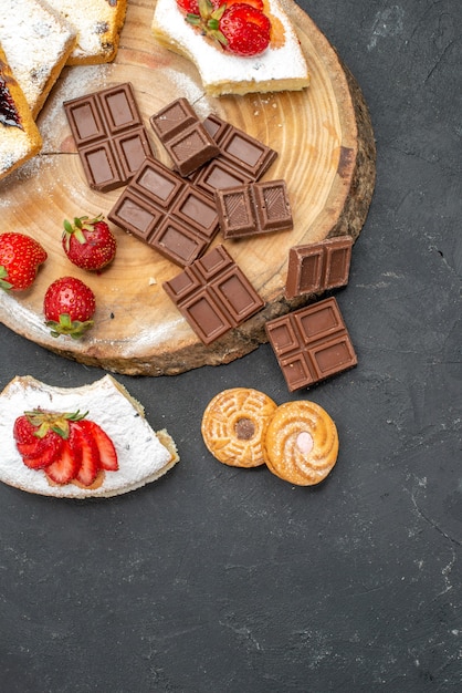 Vista superior de las rebanadas de pastel con barras de chocolate y galletas en el escritorio gris