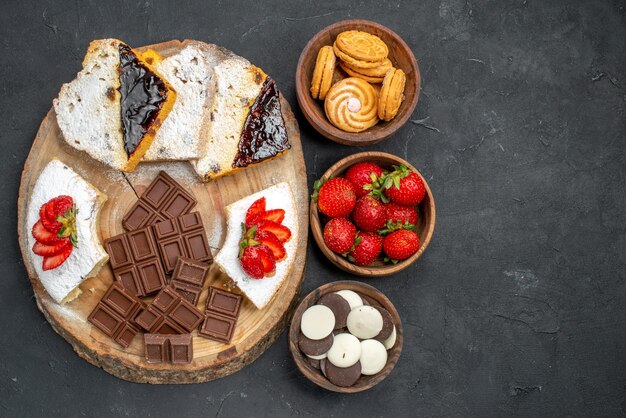 Vista superior de las rebanadas de pastel con galletas de frutas y barras de chocolate en una superficie oscura