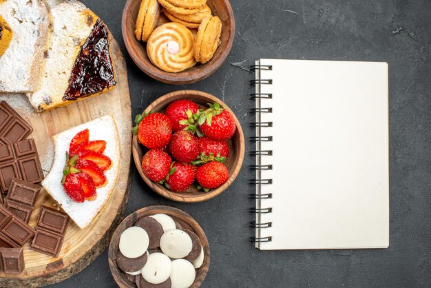 Vista superior de las rebanadas de pastel con galletas de frutas y barras de chocolate en una superficie oscura