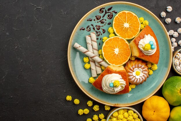 Vista superior de rebanadas de pastel con mandarinas frescas en rodajas y dulces en el escritorio oscuro