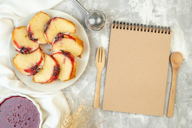 Vista superior rebanadas de pastel de mermelada en un plato mermelada en un tazón cuchara de madera y tenedor cuaderno en la mesa