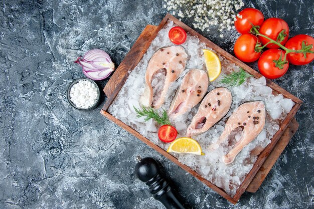 Vista superior rebanadas de pescado crudo con hielo en tablero de madera tomates cebolla sal marina en mesa