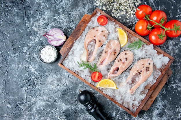 Vista superior rebanadas de pescado crudo con hielo en tablero de madera tomates cebolla sal marina en mesa