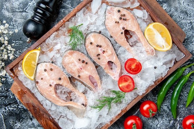 Vista superior rebanadas de pescado crudo con hielo en tablero de madera tomates molinillo de pimienta en la mesa