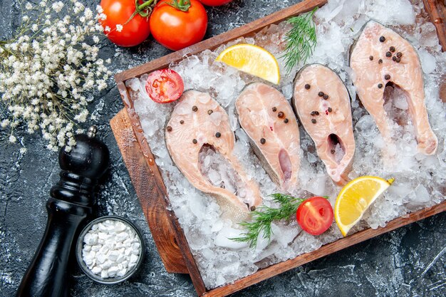 Vista superior rebanadas de pescado crudo con hielo en tablero de madera tomates sal marina en tazón pequeño molinillo de pimienta en la mesa