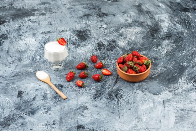 Vista superior de un recipiente de vidrio de yogur en mantel de mimbre con cuchara de madera y fresas en mármol azul oscuro y superficie de tablero de madera blanca. vertical