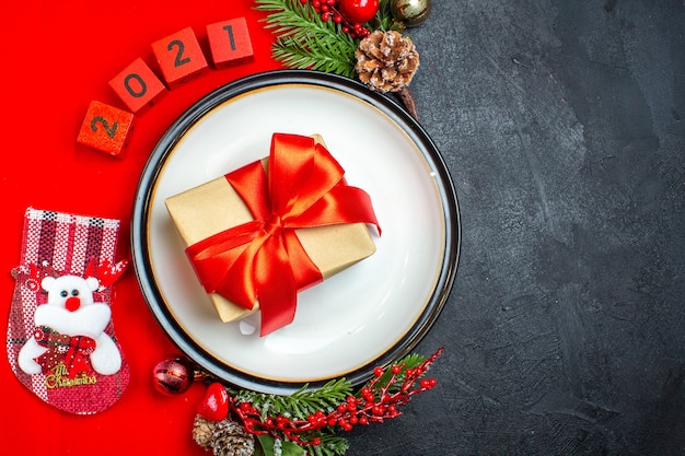 Vista superior del regalo con cinta en accesorios de decoración de plato de cena ramas de abeto y números calcetín de navidad en una servilleta roja sobre fondo negro