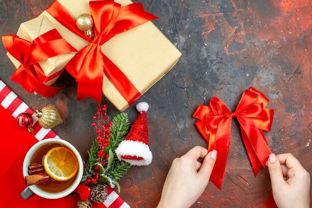 Vista superior de regalos de navidad atados con cinta roja sombrero de santa lazo rojo en mano femenina taza de té en la mesa de color rojo oscuro