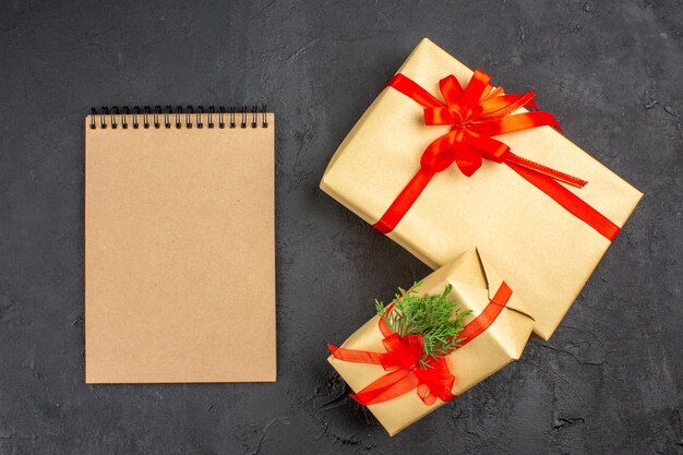 Vista superior de regalos de Navidad grandes y pequeños en papel marrón atado con cinta roja un cuaderno sobre fondo oscuro