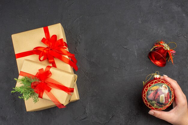 Vista superior de regalos de Navidad en papel marrón atado con cinta roja juguete de árbol de Navidad en mano femenina sobre fondo oscuro