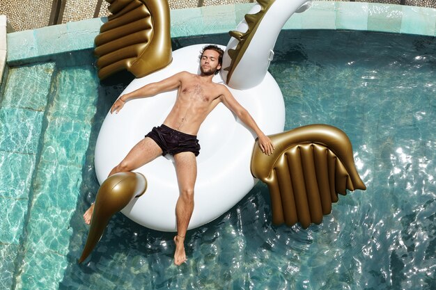 Vista superior del relajado y feliz joven descamisado flotando en la piscina, acostado en la cama de aire durante sus tan esperadas vacaciones en un país tropical.