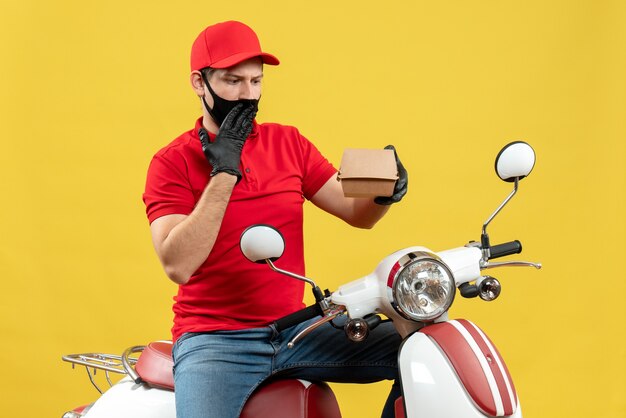 Vista superior del repartidor confundido sorprendido con uniforme y guantes en máscara médica sentado en scooter mostrando orden