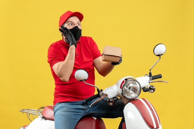 Vista superior del repartidor confundido con uniforme y guantes de sombrero en máscara médica sentado en scooter mostrando orden