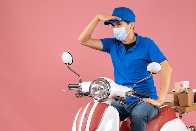 Vista superior del repartidor enfocado en máscara médica con sombrero sentado en scooter sobre fondo melocotón pastel