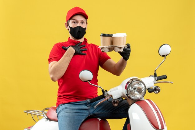 Vista superior del repartidor incierto con uniforme y guantes de sombrero en máscara médica sentado en scooter mostrando órdenes apuntando a sí mismo