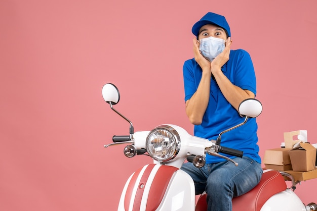 Vista superior del repartidor en máscara médica con sombrero sentado en scooter y sentirse sorprendido sobre fondo de melocotón pastel