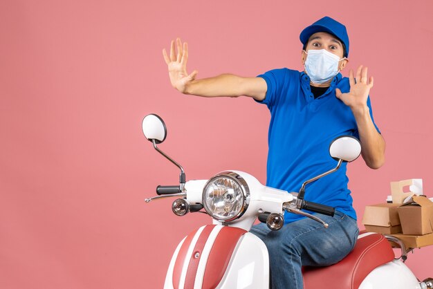 Vista superior del repartidor con máscara médica con sombrero sentado en scooter y sintiéndose asustado sobre fondo melocotón pastel