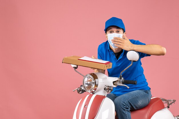 Vista superior del repartidor masculino interesado en máscara con sombrero sentado en scooter entregando pedidos con documento sobre fondo melocotón