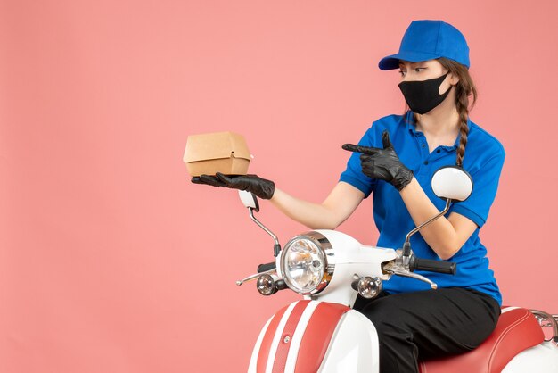 Vista superior del repartidor ocupado con máscara médica y guantes sentado en scooter entregando pedidos sobre fondo melocotón pastel